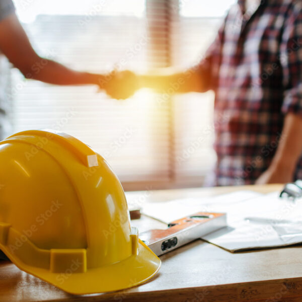 yellow safety helmet on workplace desk with construction worker team hands shaking greeting start up plan new project contract in office center at construction site, partnership and contractor concept